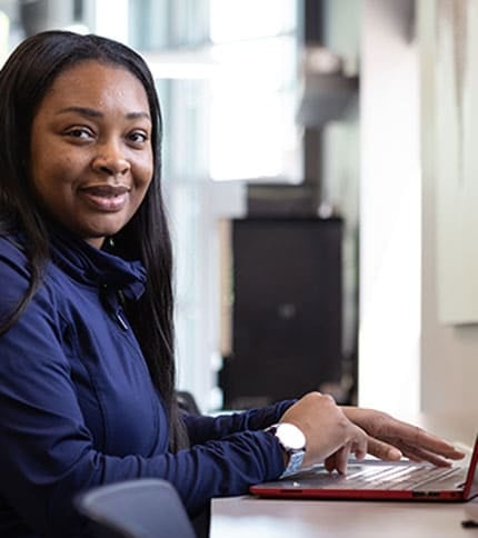 A person smiling while sitting at a desk, typing on a laptop. They're wearing a dark jacket and a watch, with a bright, modern office environment in the background.