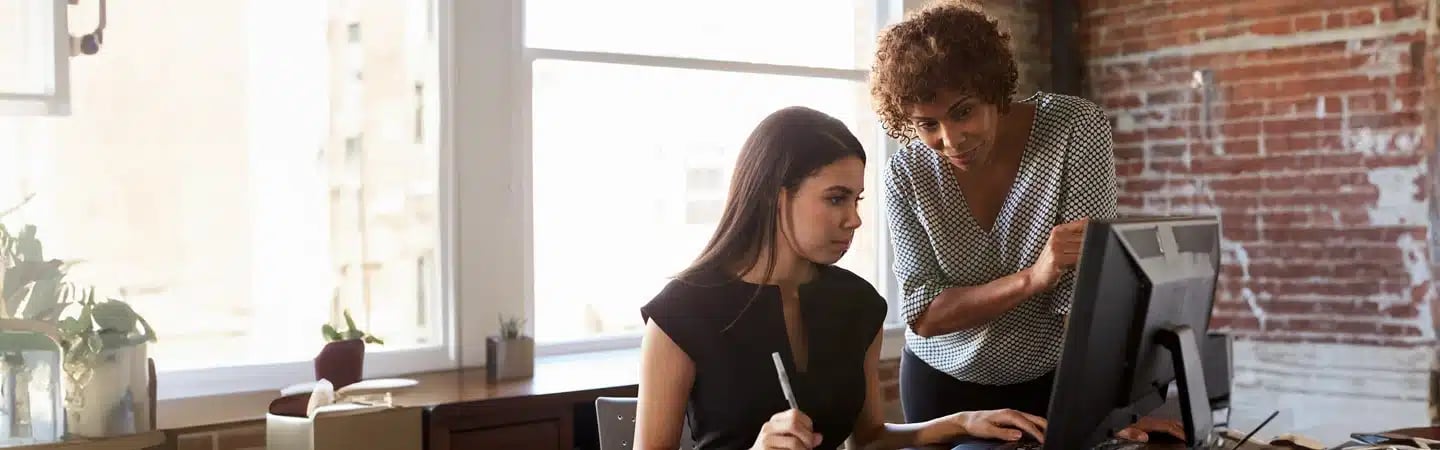 Two women work together at a desk with a computer in a well-lit office with brick walls.