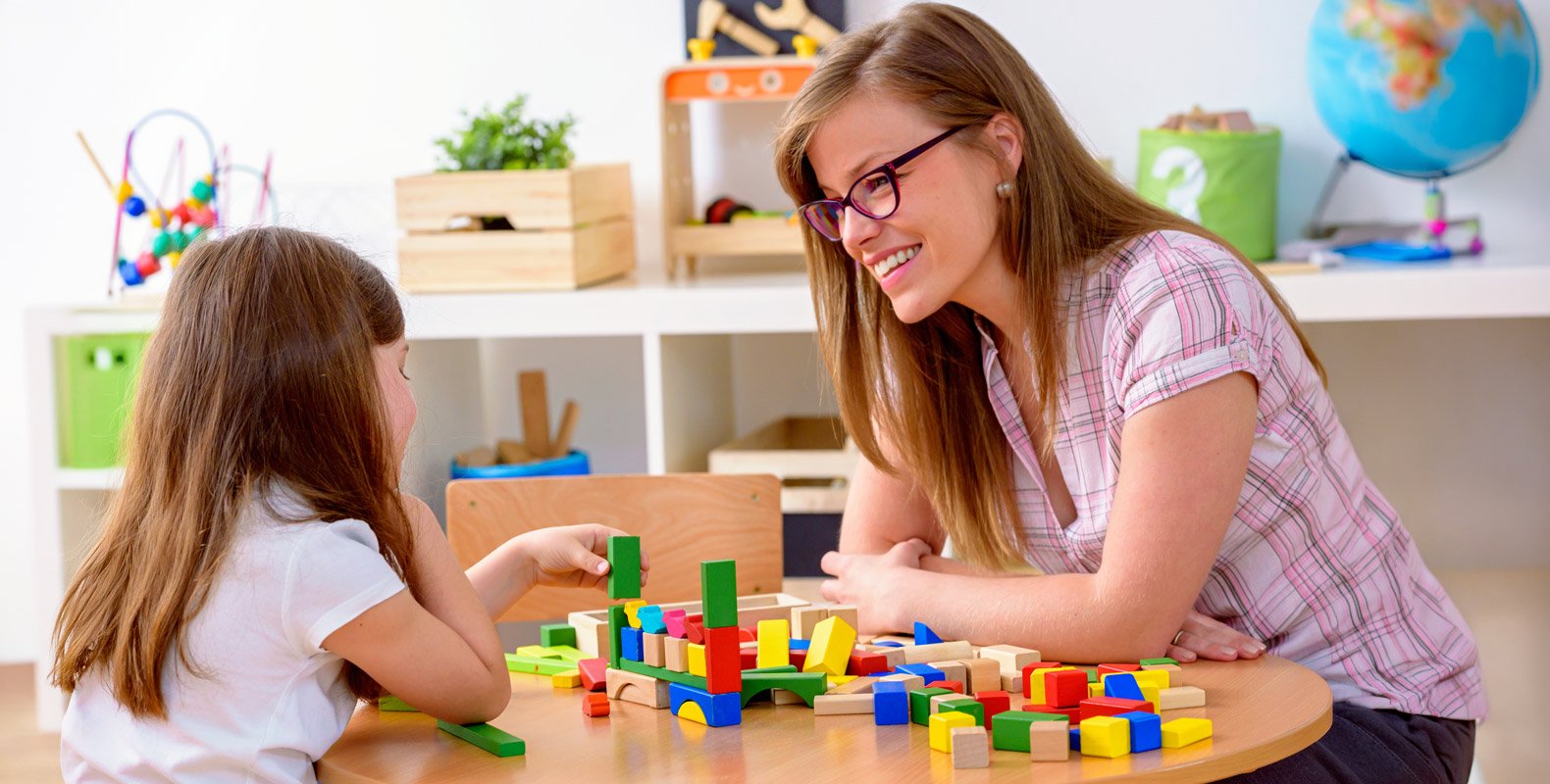 A woman and a young girl sit at a table playing with colorful wooden blocks. The woman, pursuing a childcare career, smiles at the girl in a bright, organized classroom filled with educational toys and a globe in the background.
