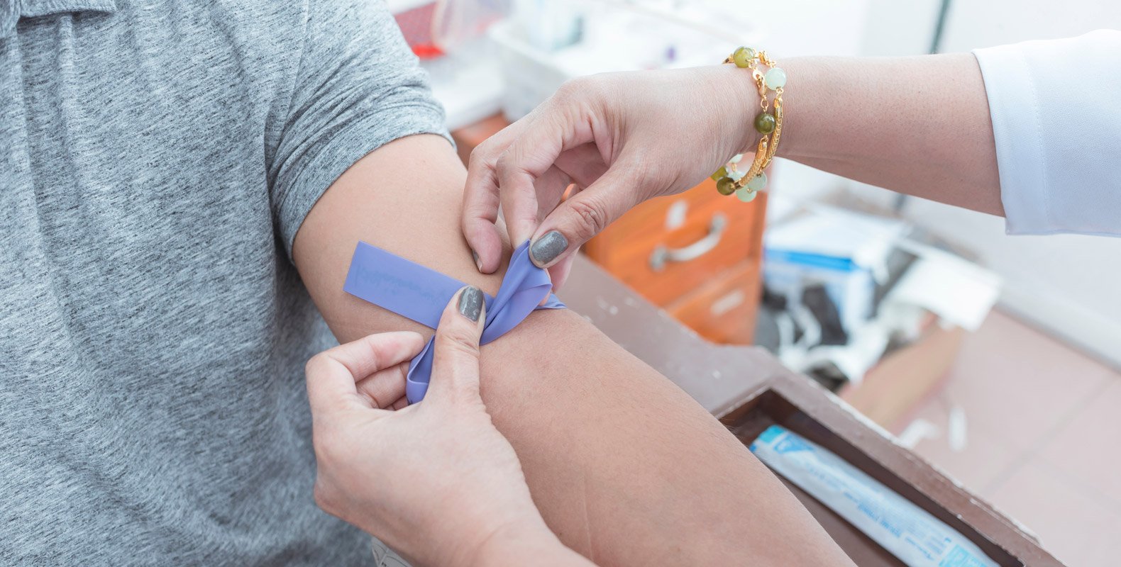 A person wearing a gray shirt is having a tourniquet tied on their upper arm by someone preparing for a medical procedure, likely a blood draw, in a clinical setting.