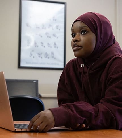 A person wearing a maroon hijab and hoodie sits at a desk using a laptop. They appear focused, with a poster visible on the wall in the background.
