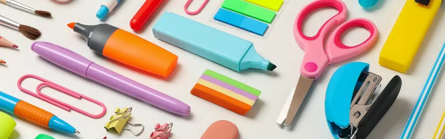 An assortment of colorful office and school supplies, including pens, scissors, sticky notes, and a stapler, on a white surface.