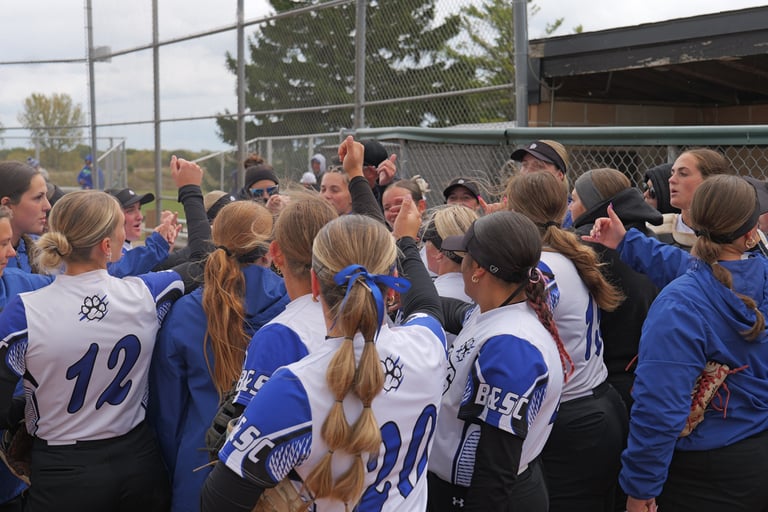 A girls’ softball team stands in a circle, arms raised together in a huddle, wearing blue and white uniforms near a fenced dugout on a cloudy day—united with the same spirit that drives students to enroll at Bryant & Stratton College for the 2026 Fall Semester.