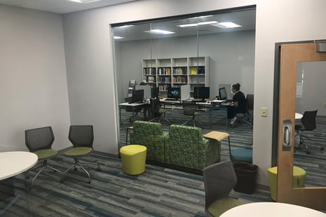 A small office space with glass walls, equipped with desks, computers, and a single person working inside.