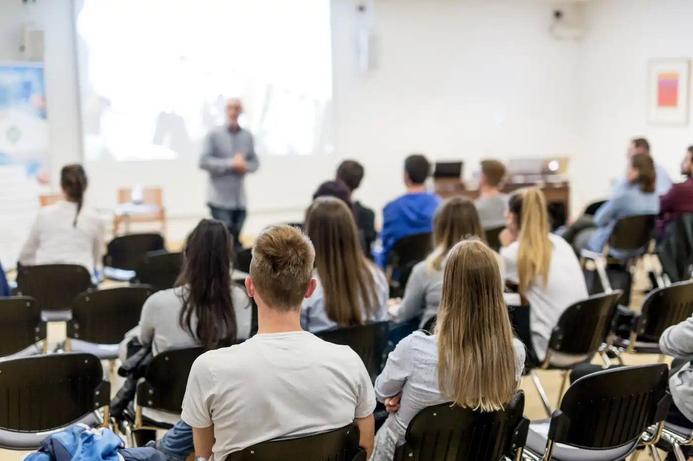 A group of people sitting in chairs facing a person giving a presentation in a brightly lit room.