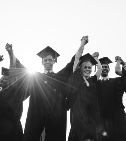 A group of graduates in caps and gowns stand together outdoors, smiling and holding hands raised in celebration, with sunlight shining behind them.