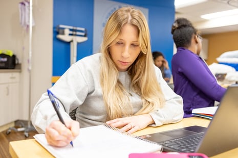 Woman writing in a notebook at a table with a laptop nearby; others are working in the background.