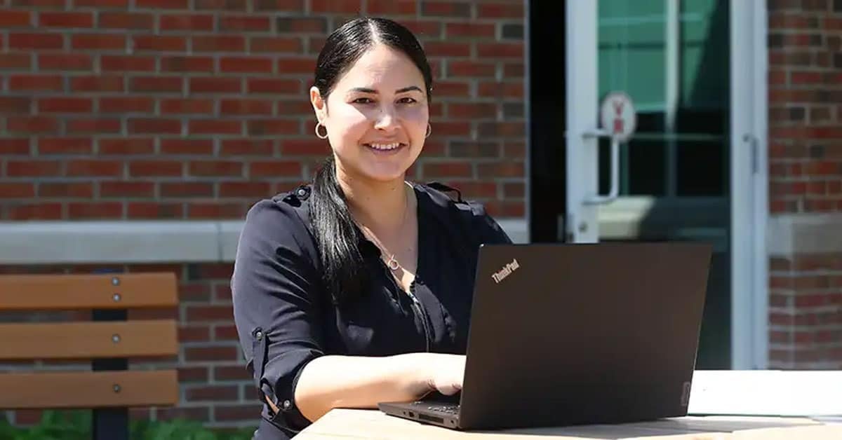 A woman with dark hair smiles while working on a laptop at an outdoor table near a brick building.