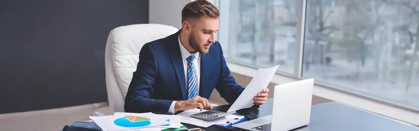 Man in a suit working at a desk with a laptop, calculator, and papers. He is looking at a document thoughtfully.