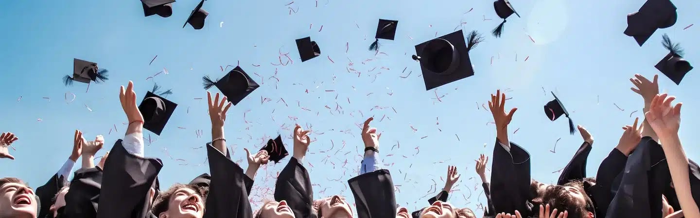Graduates in caps and gowns celebrating, throwing their graduation caps into the air against a clear blue sky.