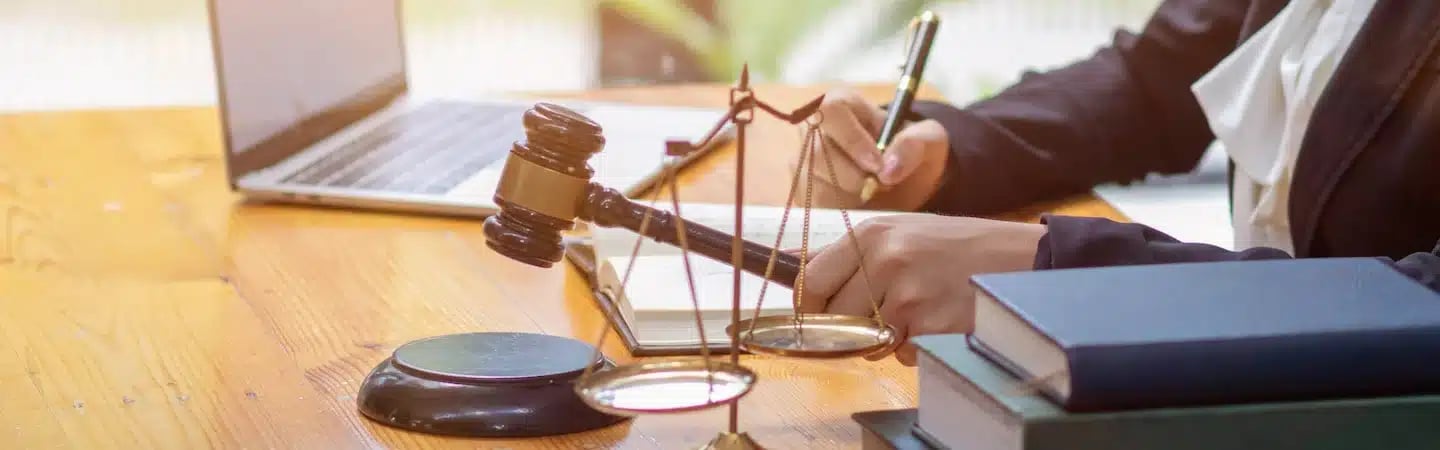Person writing at a desk with a gavel, scales of justice, laptop, and books.
