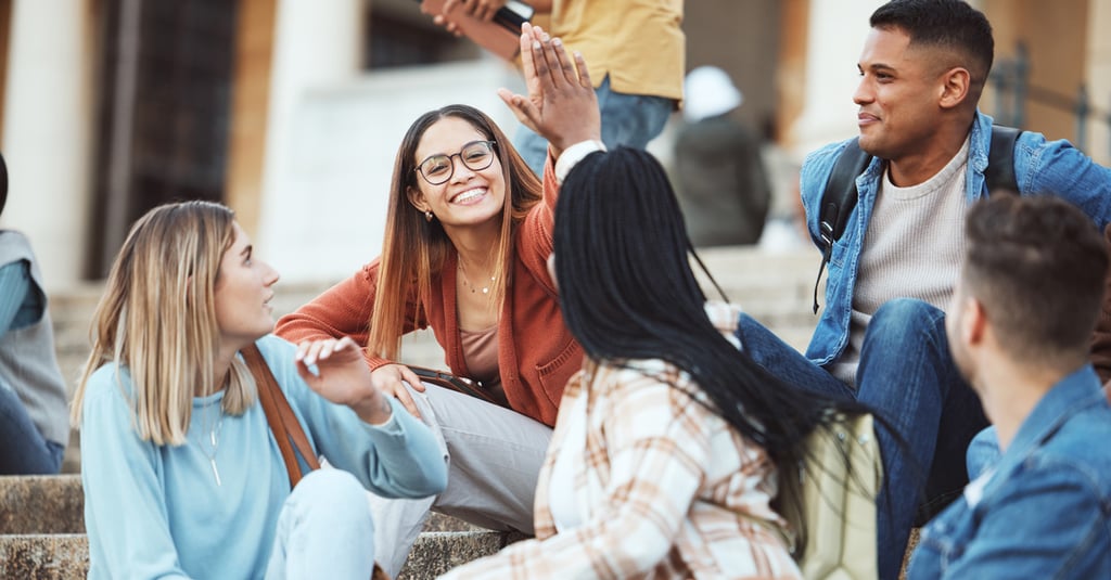 A group of five young adults sit on outdoor steps, smiling and talking. Two people in the center share a high five, celebrating good news about Financial Aid and creating a cheerful, friendly atmosphere.