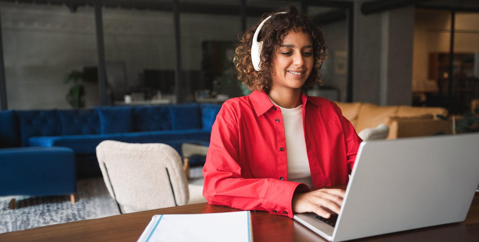 A person wearing headphones and a red shirt smiles while typing on a laptop at a desk, with a notebook nearby and modern office furniture in the background.