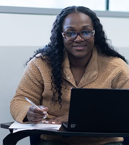 A person with long hair and glasses is smiling while sitting at a desk, working on a laptop with papers nearby. They are wearing a tan fleece jacket, perhaps studying for their health services administration associate degree. The background shows a light-colored wall and a window.