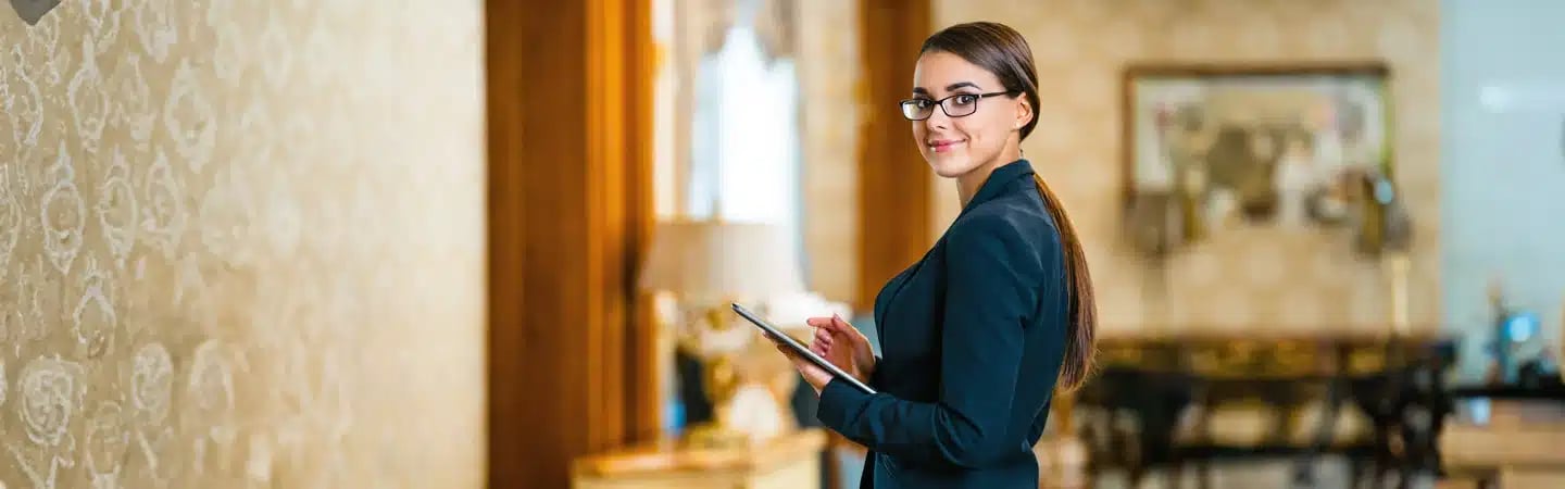 A woman in a suit and glasses holds a tablet, standing in an elegantly decorated room with ornate wallpaper.
