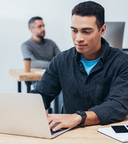 A young man wearing a dark shirt works on a laptop at a desk, possibly researching FAQs or terminology, while another person sits blurred in the background. A smartphone and coffee cup are on the desk.