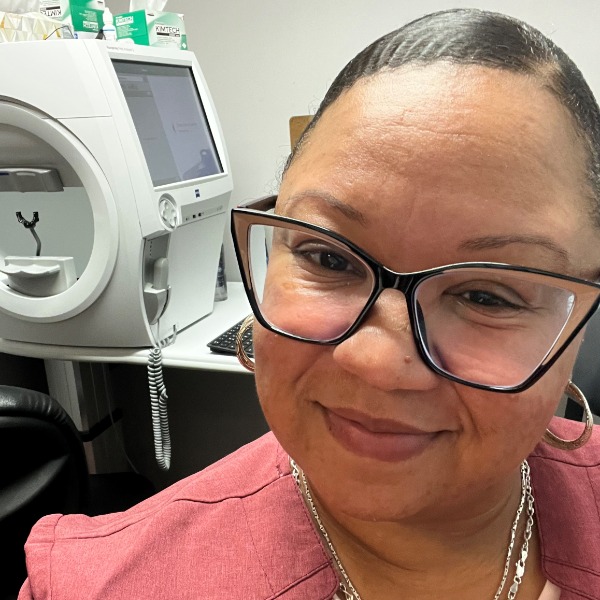A woman wearing glasses and a pink blazer smiles at the camera in an Albany office setting, with medical equipment and supplies visible in the background; she is identified as Asya Mayotte.