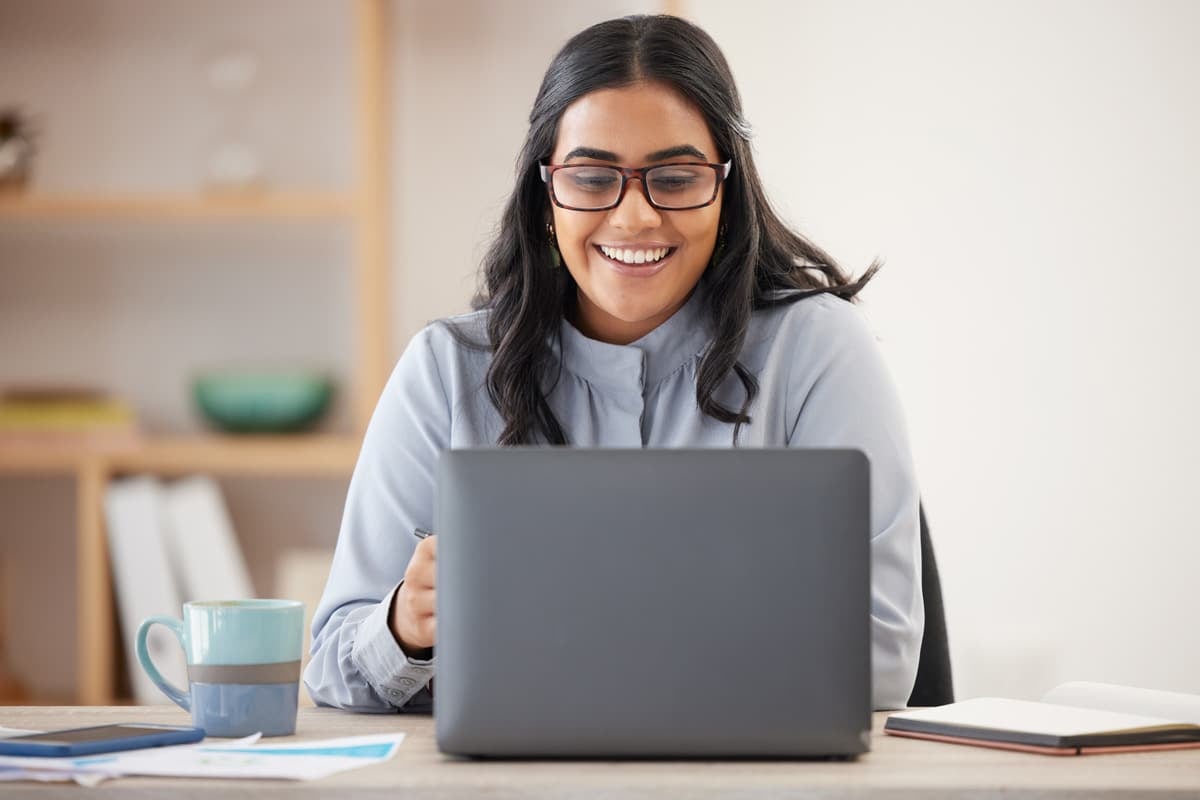 Smiling person with glasses working on a laptop at a desk with a mug and papers in a home office.