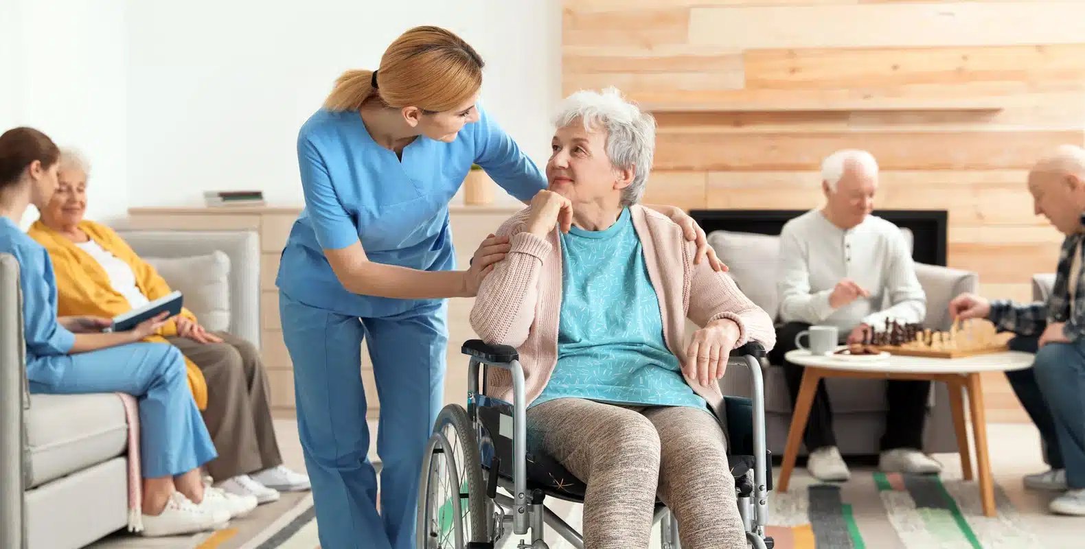 A caregiver in blue scrubs talks to an elderly woman in a wheelchair; other seniors are engaging in activities nearby.