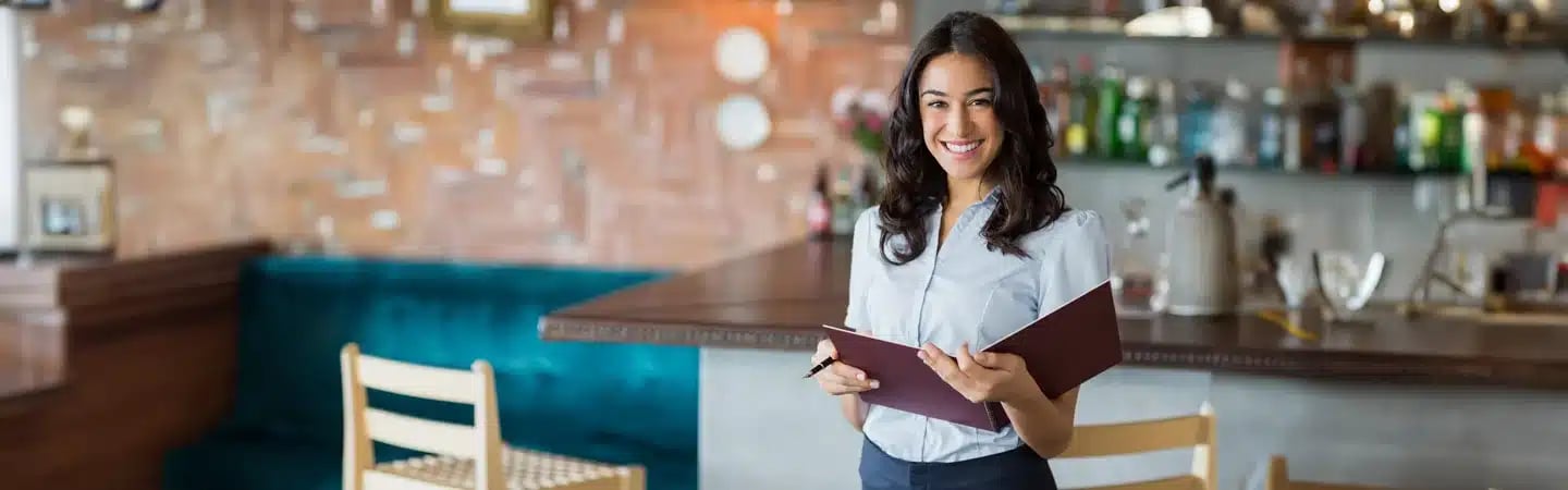 A smiling woman holding a menu stands in a stylish restaurant with a bar and wooden chairs in the background.