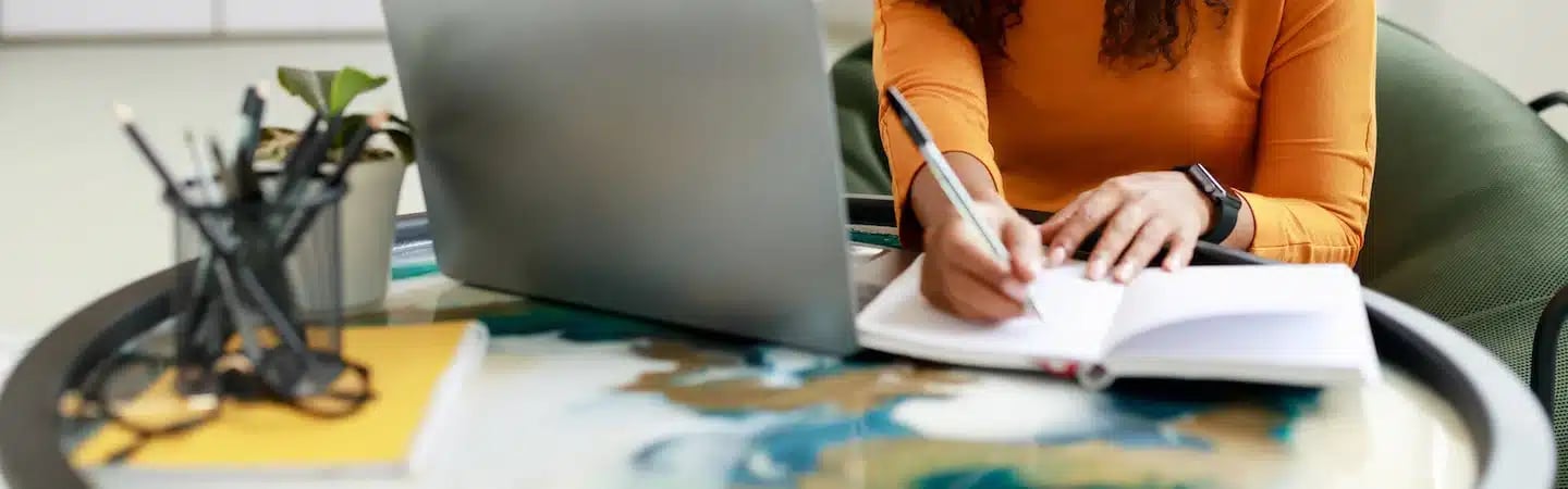 Person in yellow shirt writing in a notebook at a desk with a laptop, plant, and stationery holder.