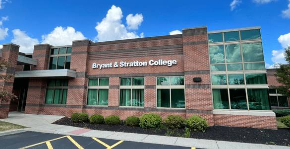 A red brick building with large windows and a sign that reads "Bryant & Stratton College" against a blue sky with clouds.