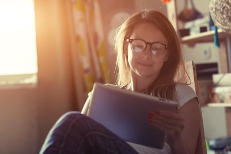 A woman with glasses sits by the window, reading a tablet with a smile in a cozy, sunlit room.