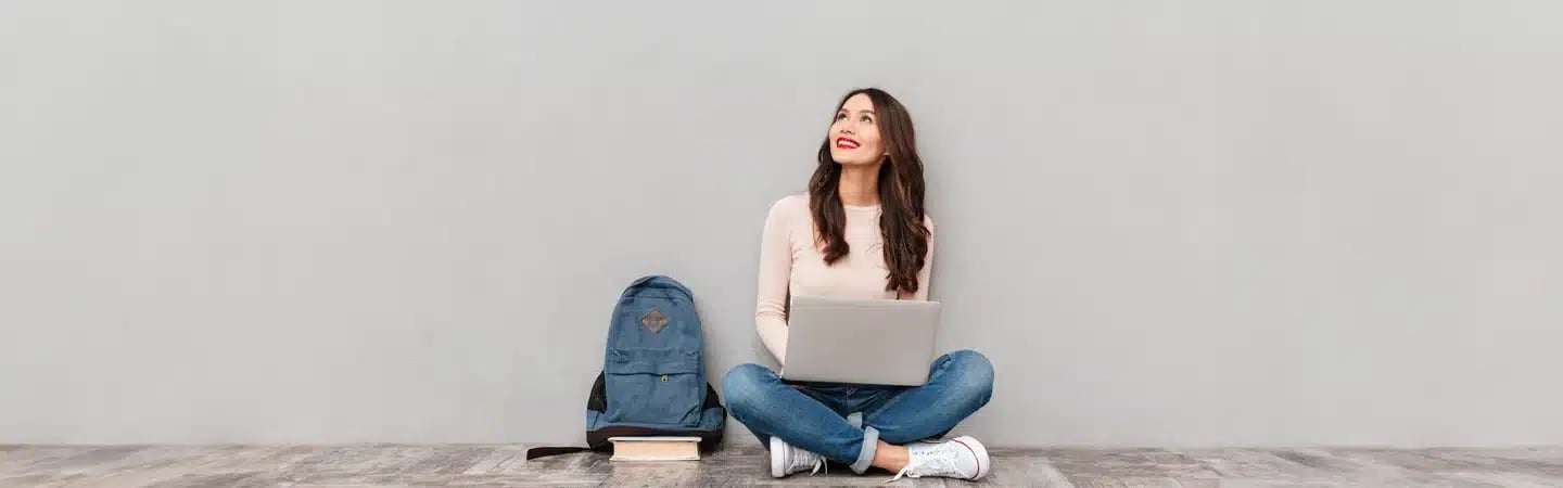 A woman sits cross-legged on the floor with a laptop, smiling and looking up. A backpack and books are next to her.