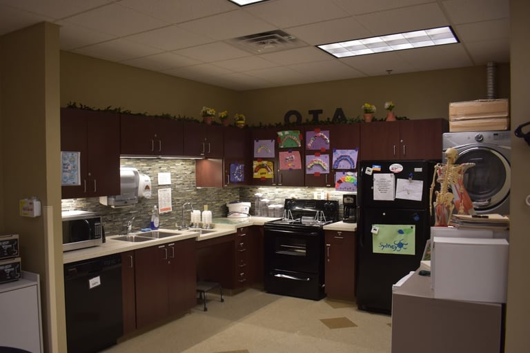 A kitchenette with dark wood cabinets, a sink, stove, refrigerator, dishwasher, and children's artwork on display.