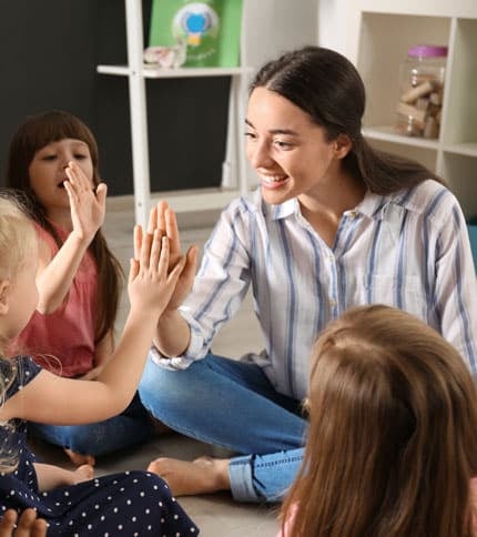 A smiling woman with an Early Childhood Certificate sits cross-legged on the floor, high-fiving a young girl. Two other children sit nearby, watching and smiling in admiration. They are in a room with shelves holding various items in the background.