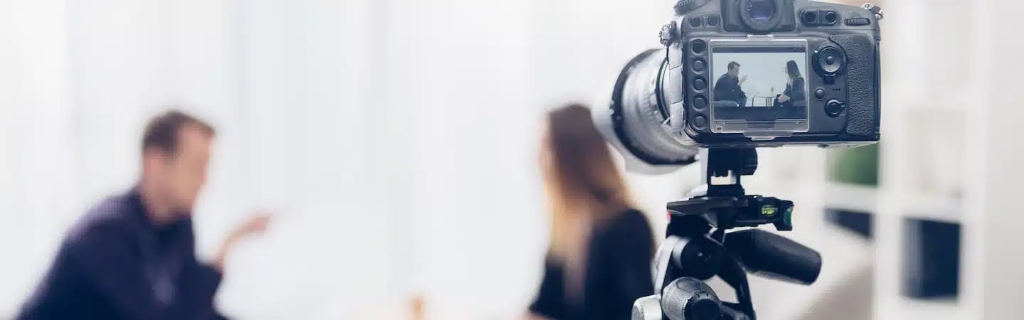 Close-up of a camera recording an interview, with two blurred people sitting and talking in the background.