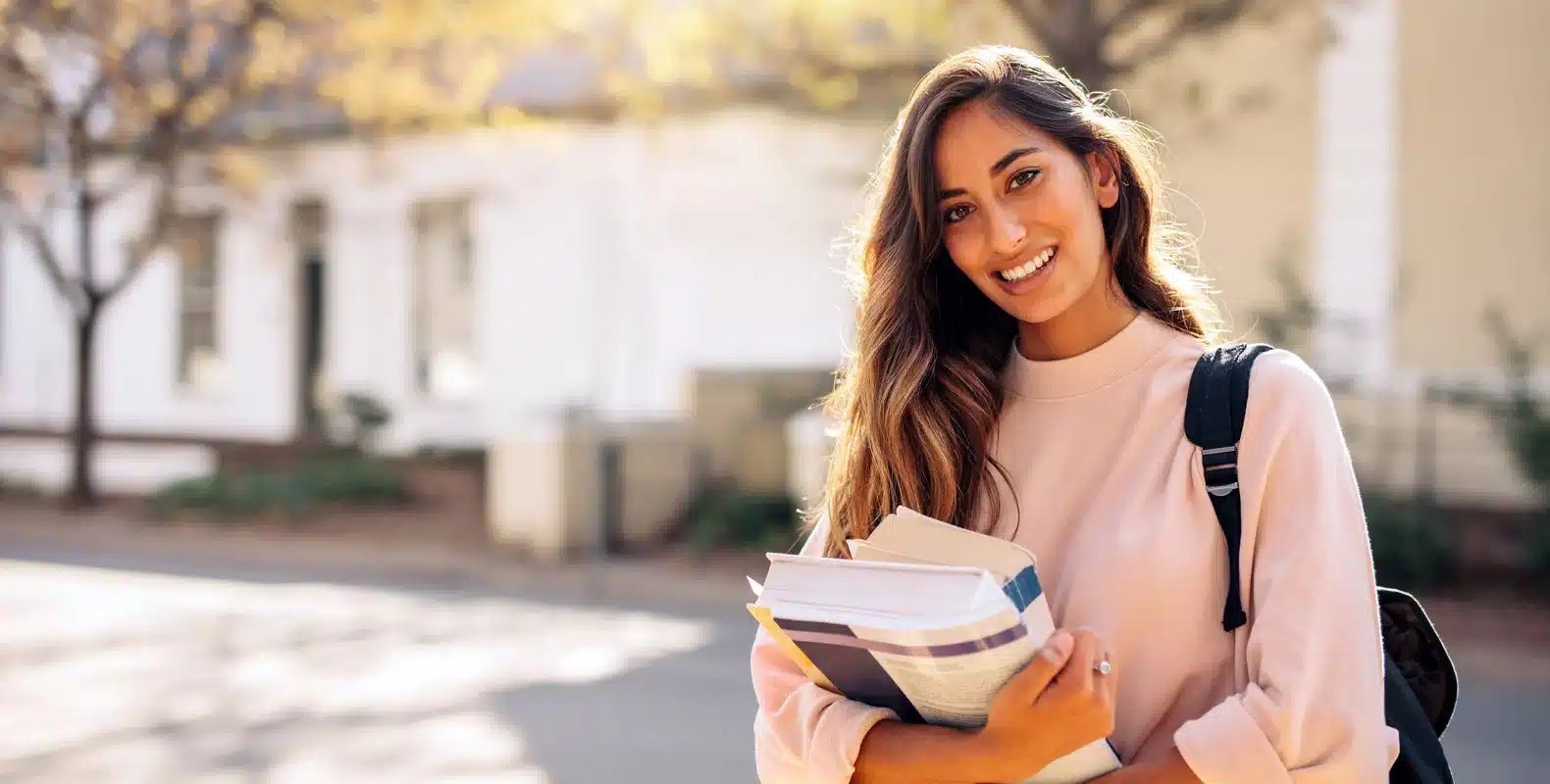 A smiling woman holding books and a backpack stands outdoors on a sunny day.