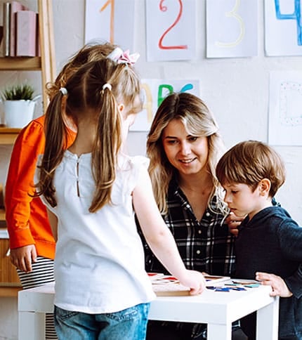 A woman sits at a table with three children engaged in an activity. They appear to be focused on something in front of them, with colorful papers and numbers on the wall in the background, suggesting a learning environment.