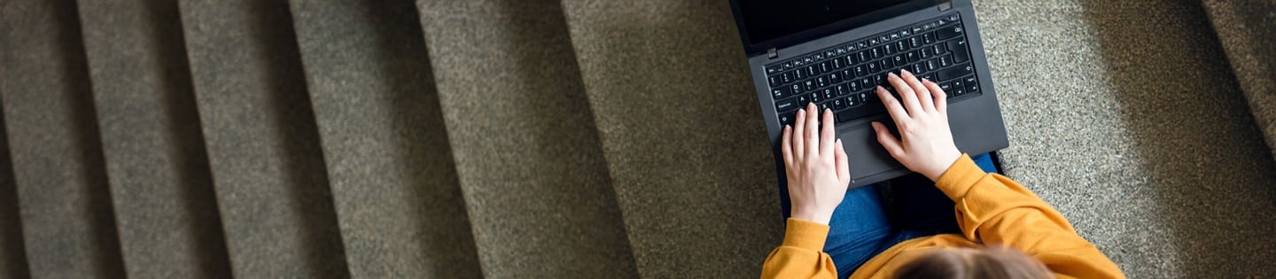 Person in a yellow sweater using a laptop while sitting on carpeted stairs, viewed from above.