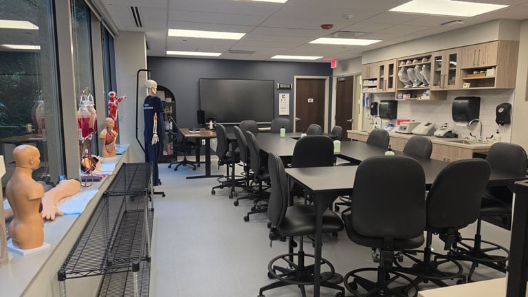 Medical assisting lab with exam tables, rolling chairs, cabinets, sinks, supplies, and training mannequins for hands-on practice.