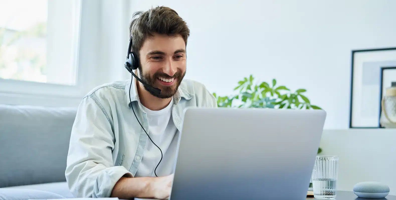 Man wearing a headset, smiling while using a laptop at a desk, with a plant and glass of water in the background.