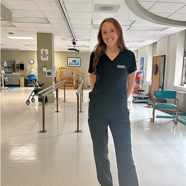 A person stands smiling in a medical uniform in a brightly lit, clean hospital or clinic setting.
