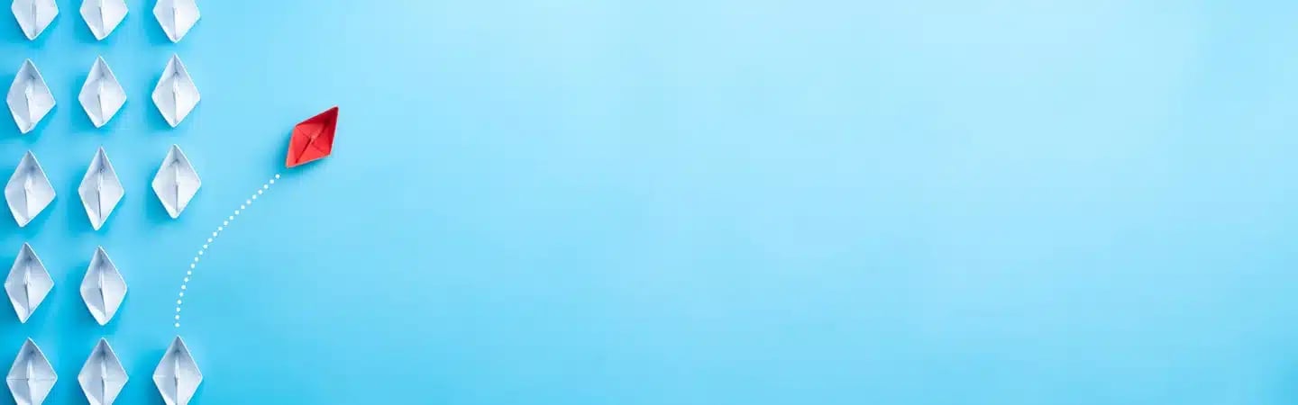 A red paper boat diverts from a group of white paper boats, symbolizing individuality and leadership on a light blue background.