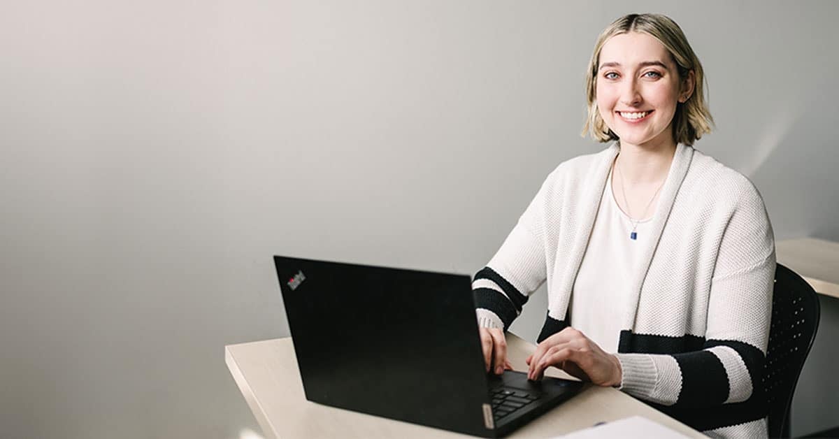 A person with short hair and striped sweater smiles while typing on a laptop at a desk.