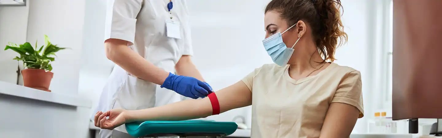A healthcare worker prepares to draw blood from a masked woman sitting in a medical clinic.