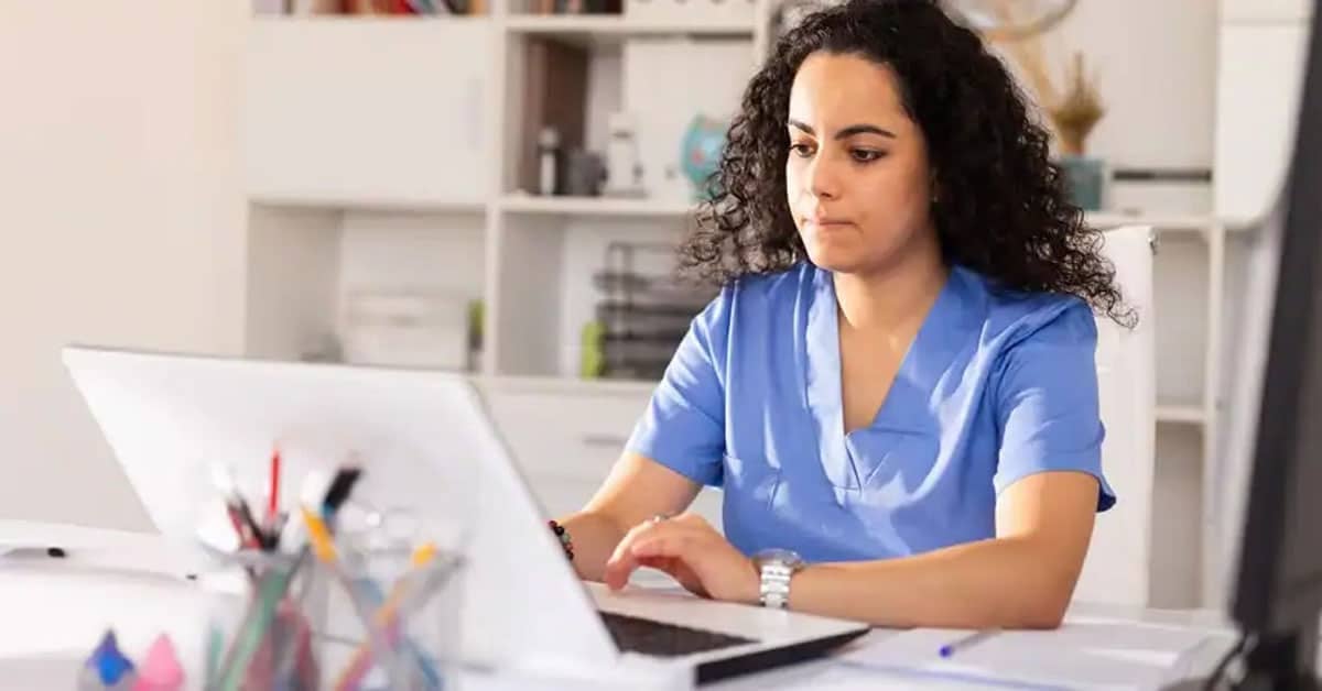 A person with curly hair wearing scrubs uses a laptop at a desk in an office setting.