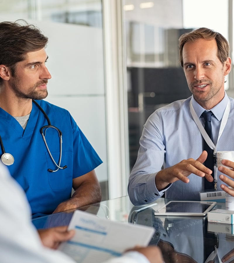 Two male healthcare professionals, including a Patient Care Coordinator in blue scrubs and a colleague in a dress shirt with a lanyard, sit at a table discussing paperwork with their team in a modern office setting.