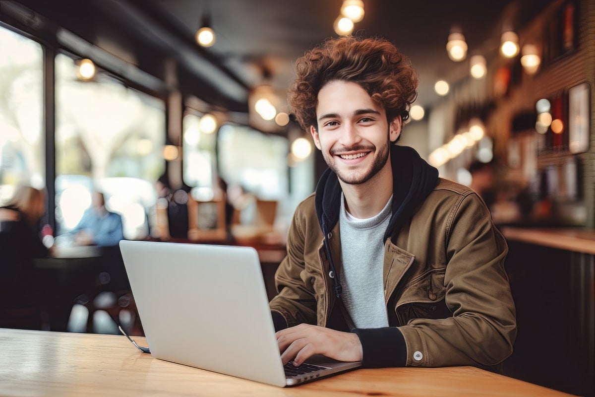 A smiling young man with curly hair works on a laptop in a cozy café with warm lighting and blurred background.