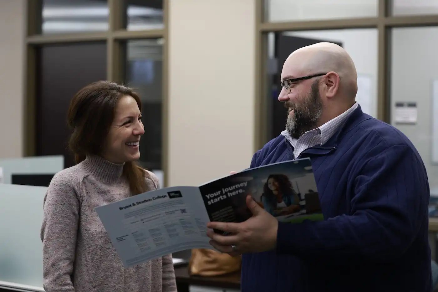 Two people are smiling and discussing a brochure in an office setting.