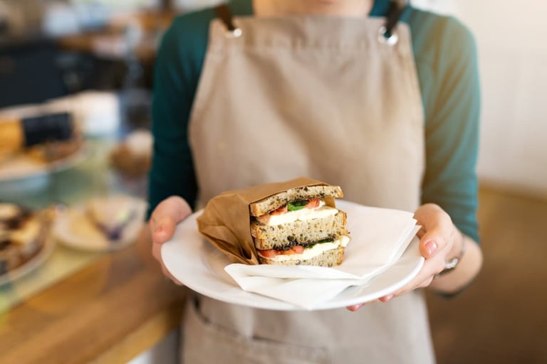 Person in a brown apron holding a white plate with a wrapped sandwich on it.