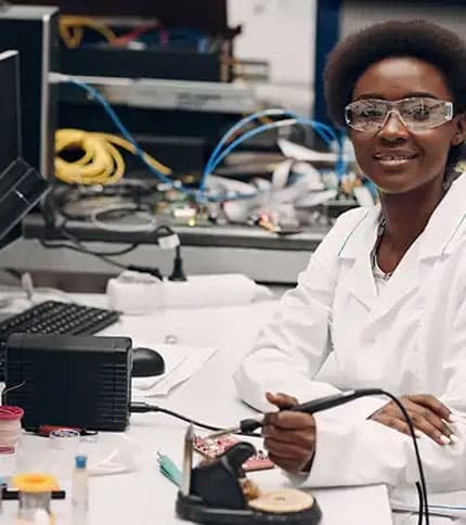 A scientist in a lab coat and safety goggles sits at a desk with electronic equipment and tools. She holds a soldering iron and smiles at the camera. Various wires and lab items are visible in the background.
