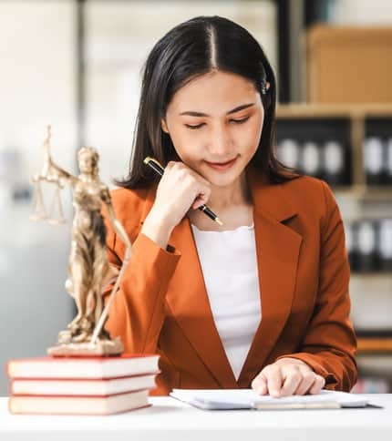 A woman in an orange blazer studies a document at a desk, resting her chin on one hand. In front of her are stacked books and a bronze statue of Lady Justice. Shelves with files are visible in the background.