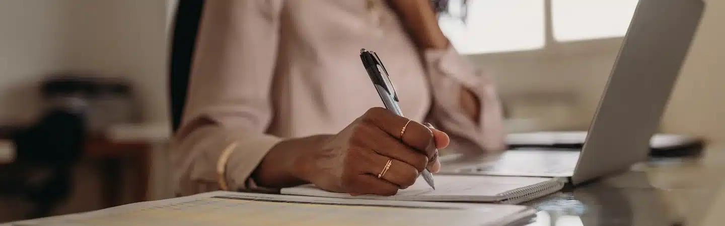 Person wearing a light shirt writes in a notebook with a pen next to a laptop on a desk.