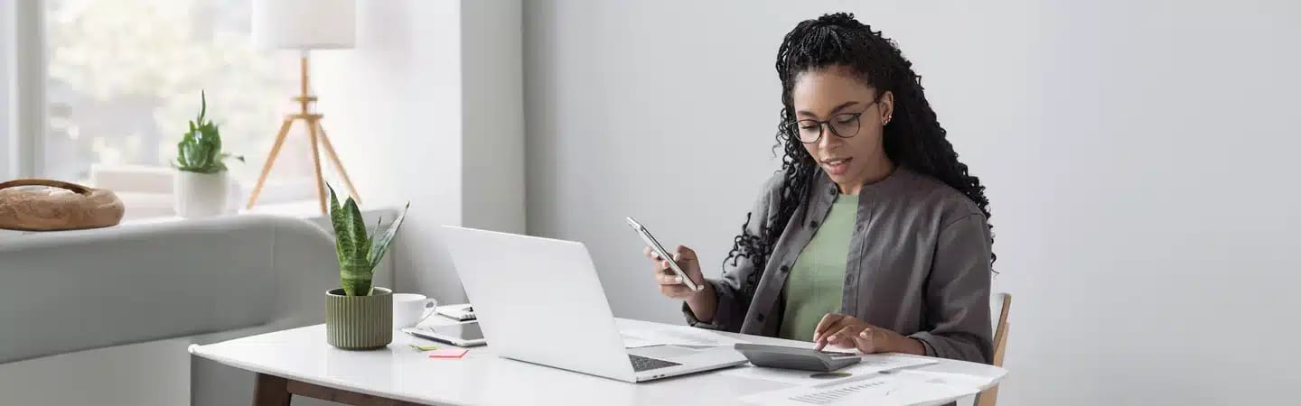 Woman working at a desk with a laptop, calculator, and papers, holding a smartphone, plants and decor around her.