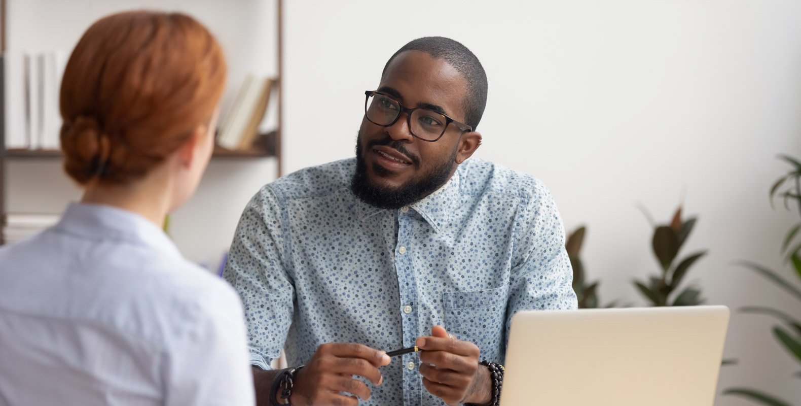 A man with glasses and a beard sits at a desk with a laptop, discussing Human Services Careers with a red-haired woman in an office setting filled with bookshelves and plants.